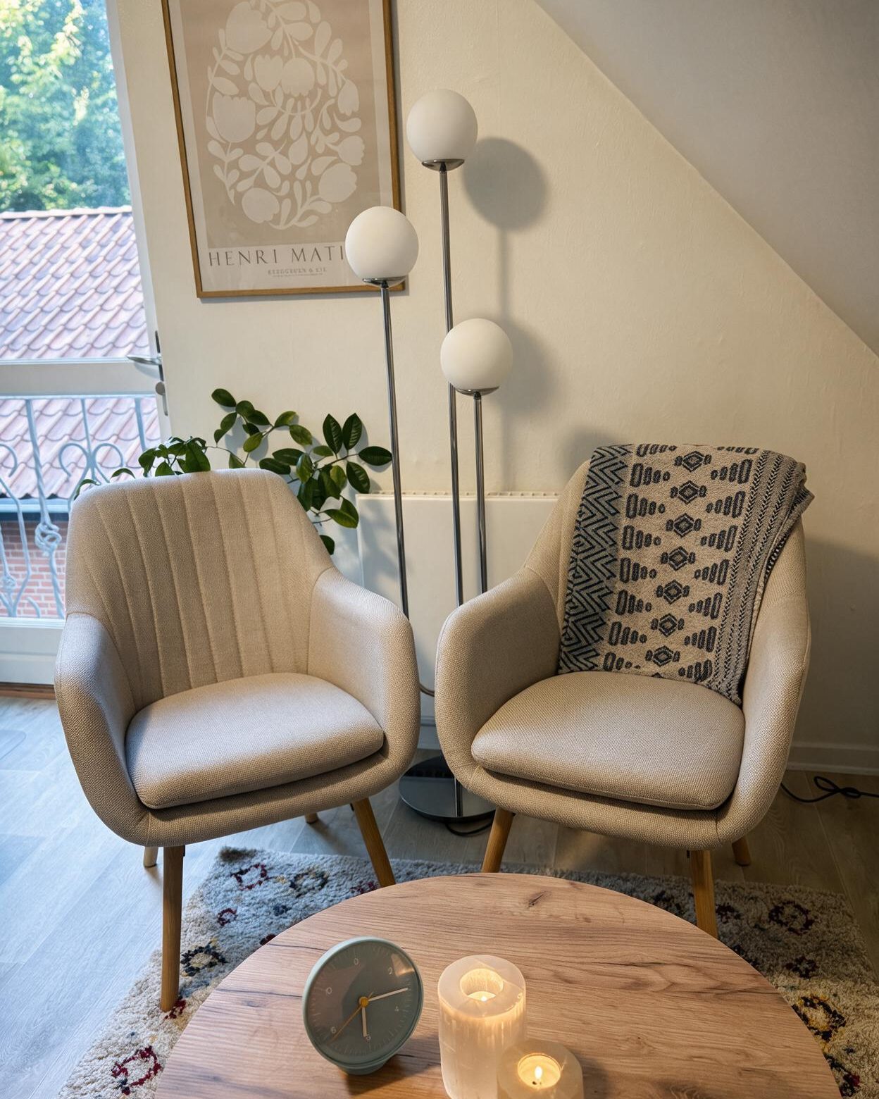 To lyse lænestole side om side under skråvæggen, med en gulvlampe, en plakat og et rundt egetræsbord med tændte lys i forgrunden